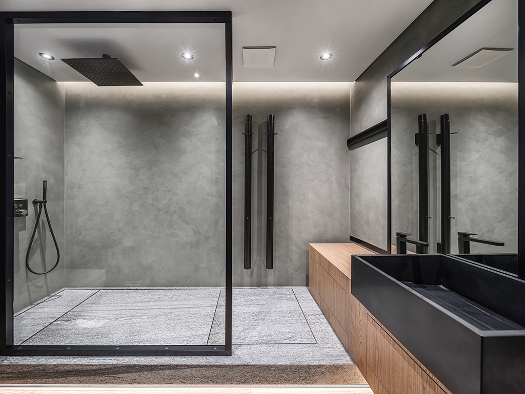 Bathroom in a modern style with gray tiled walls. There is a shower with a glass partition, wooden stand with a black sink and a faucet, large mirror, luminous lamps. Horizontal.